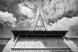 Schwarz-Weiß-Fotografie eines Billboards mit dreieckigem Aufbau über einem Blechdach, dramatischer Himmel im Hintergrund. Architekturdetail von J.F. Lindemann, Leica M, Summicron 28mm ASPH