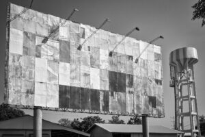 Schwarz-Weiß-Fotografie einer überklebten Plakatwand beim Flughafen Chiang Rai in Thailand, daneben ein Wasserturm – Teil der Serie Billboards von J.F. Lindemann, Leica M und Summicron 28mm ASPH