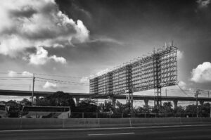 Schwarz-Weiß-Fotografie eines riesigen leeren Billboard-Gerüsts an einer Schnellstraße in Thailand, im Hintergrund Hochstraße und Wolkenhimmel – Teil der Serie Billboards von J.F. Lindemann, Leica M und Summicron 28mm ASPH