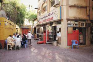 Straßenszene in Dubai: Männer sitzen vor dem Maaraj Restaurant auf bunten Monobloc-Stühlen, daneben ein leerer blauer Plastikstuhl – Fine Art Photography von J.F. Lindemann, aufgenommen 2014 mit Leica M und Summicron 28mm asph.