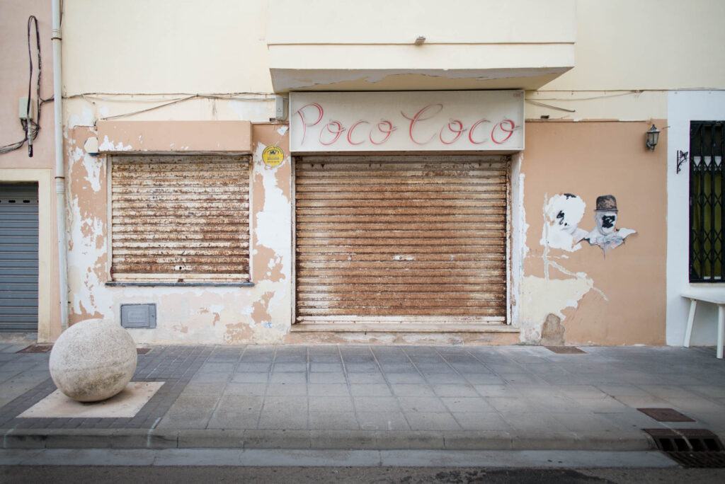 Geschlossene Ladenfront mit verblichenem Schriftzug Poco Coco und rostigen Rolltoren in Spanien – Fotografie aus der Serie FERMÉ-10 von J.F. Lindemann mit Leica M und Summicron 28mm ASPH