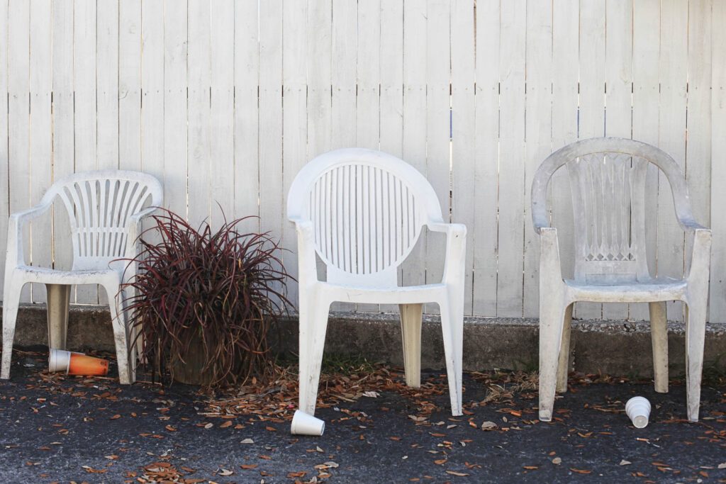 Drei gebrauchte Monobloc-Stühle vor weißem Lattenzaun, Topfpflanze in der Mitte, Herbstlaub und weggeworfene Becher am Boden – Fine Art Photography von J.F. Lindemann, Amerika 2013.