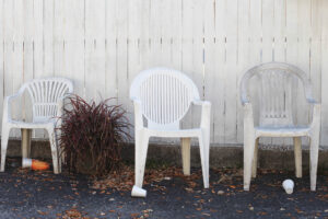 Drei gebrauchte Monobloc-Stühle vor weißem Lattenzaun, Topfpflanze in der Mitte, Herbstlaub und weggeworfene Becher am Boden – Fine Art Photography von J.F. Lindemann, Amerika 2013.