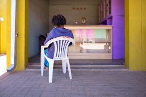 Namibia 2013: Eine Person sitzt von hinten auf einem weißen Monobloc-Stuhl vor einem kleinen Laden mit farbigen Wänden in Gelb und Violett. Fine Art Photography von J.F. Lindemann, Leica M mit Summicron 28mm ASPH.