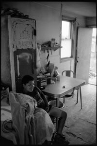 Junge sitzt in einem einfachen Wohnraum mit Tisch und Schrank, Pine Ridge Reservat, South Dakota, Schwarzweiß-Fotografie mit Leica M6 und dem Summicron 35mm von J.F. Lindemann, 1994