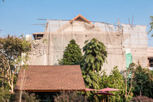 Hausfassade in Renovierung, mit Planen und Bambus Gerüsten verhüllt, im Kontrast zu tropischen Bäumen und Garten · Serie WRAPPED von J.F. Lindemann · Leica M, Summicron-M 28mm f/2.0 ASPH