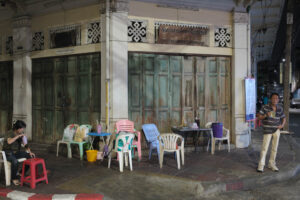 Straßenszene in Bangkok 2025: Mehrere bunte Monobloc-Stühle und ein Tisch vor einer verwitterten Holzfassade, zwei Personen im Bild. Fine Art Photography von J.F. Lindemann, Leica M mit Summicron 28mm ASPH.