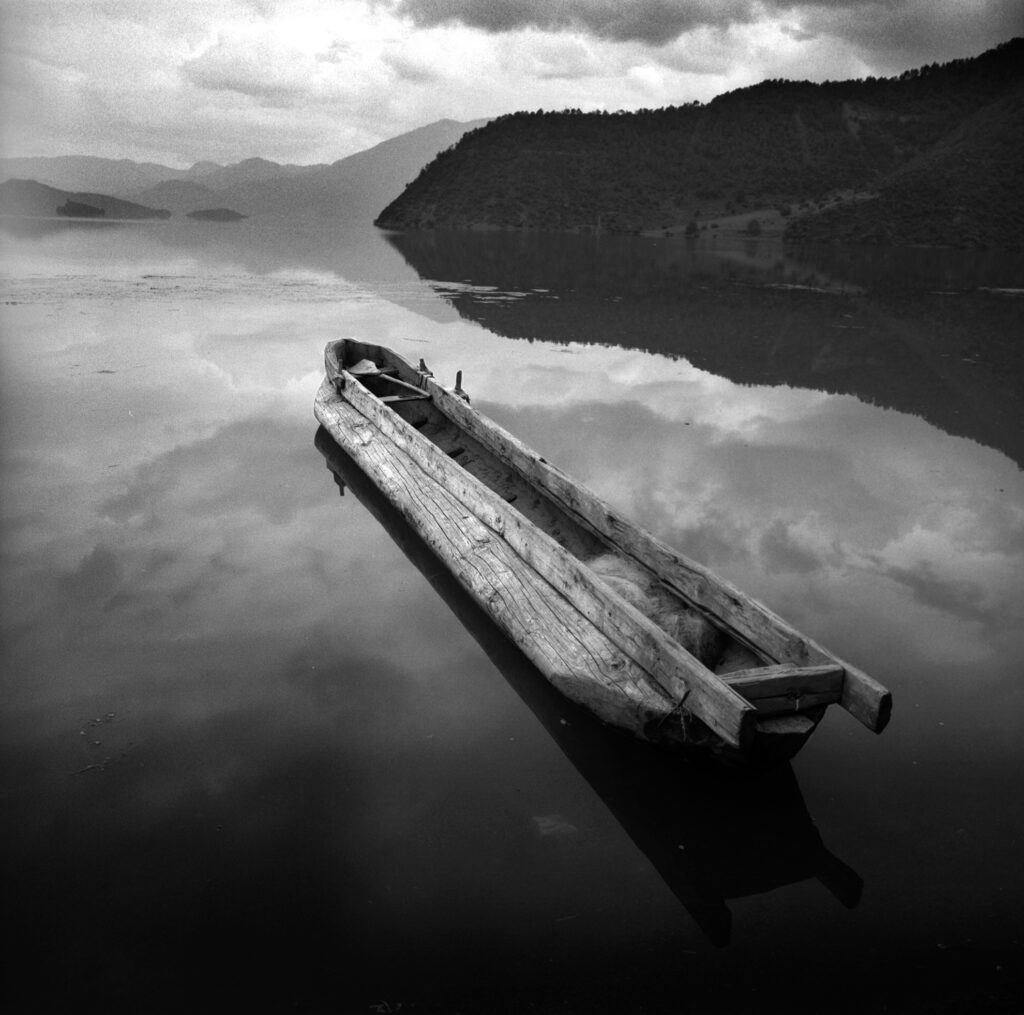 Traditionelles Holzboot auf einem stillen See, Mono Lake, in Yunnan, China, 1993, Spiegelung der Wolken im Wasser, Schwarzweiß-Fotografie mit Mamiya 6 auf Ilford von J.F. Lindemann