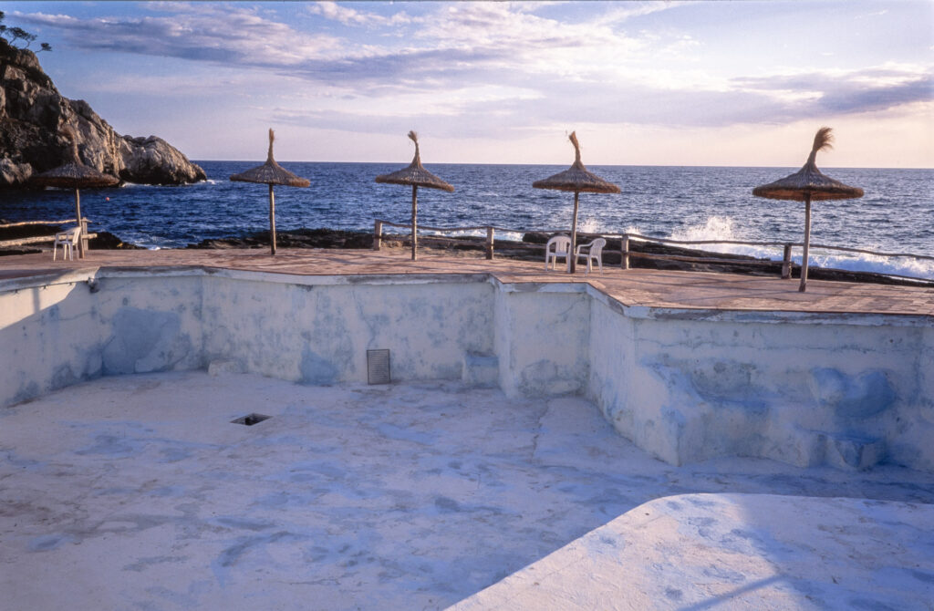 Ein leerer Pool im Vordergrund. Dahinter das Meer dazwischen 4 Sonnenschirme und ein paar monoblocs, oder nenne es Plastikstuhl.